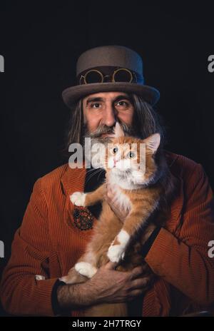 Un intellectuel à poil gris d'âge mûr dans un chapeau de steampunk pose avec un chat au gingembre. Banque D'Images