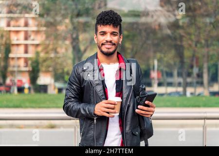 un jeune latin élégant sourit en regardant l'appareil photo avec un téléphone et un café à emporter entre ses mains, le concept de technologie et le style de vie urbain Banque D'Images