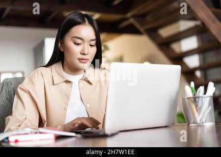 Jeune femme asiatique occupée travaillant à la maison, utilisant un ordinateur portable, assis à une table avec des livres et des crayons, tapant sur le clavier d'ordinateur Banque D'Images