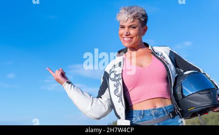 Jeune femme blonde aux cheveux courts vêtue comme un motard dans une veste de fourrure et un casque de sécurité pointe avec son doigt sur un fond bleu ciel Banque D'Images