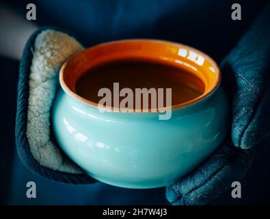 Un homme vêtu d'un tablier bleu foncé et de spéléologues tient un pot d'argile bleu ouvert avec du bouillon dans ses mains.Cuisine maison. Banque D'Images