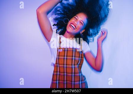Une jeune femme énergique dansant et foulant ses cheveux avec joie.Une jeune femme joyeuse riant joyeusement tout en se tenant seule dans une lumière vive au néon.Carétre Banque D'Images