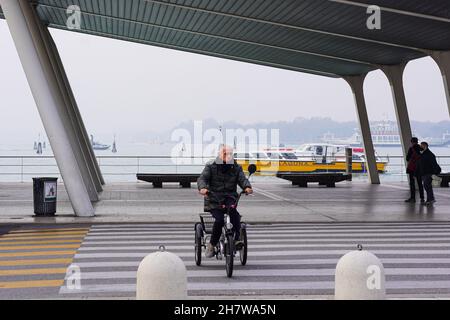 Port du Lido, île dans la lagune de Venise.en arrière-plan un bateau-bus, Vaporetto.Devant, un homme traverse un passage de côté sur sa bicyclette à trois roues. Banque D'Images