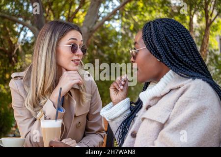 Deux jeunes amies aiment le café ensemble, assis à une table, bavarder, chuchoter et faire du grésillement.Concept de diversité et de temps de loisirs. Banque D'Images