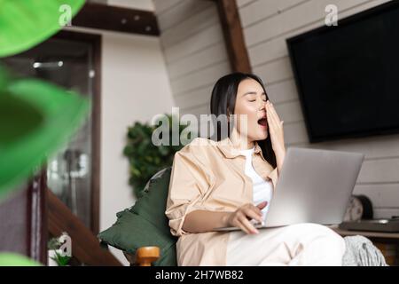 Une jeune fille asiatique fatiguée fait des sires pendant son travail sur un ordinateur portable, s'assoit à la maison, travaille à distance depuis son salon Banque D'Images