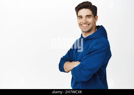 Portrait d'un jeune homme plein de sourire croisant les bras sur la poitrine, debout dans un sweat à capuche bleu, regardant déterminé à la caméra, fond blanc Banque D'Images