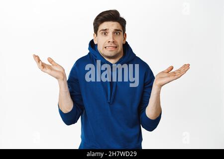 Un jeune homme confus et sans indice hausse les épaules, semble perplexe, se répand les mains sur le côté, ne sait pas, ne peut pas dire,se tient sur fond blanc Banque D'Images