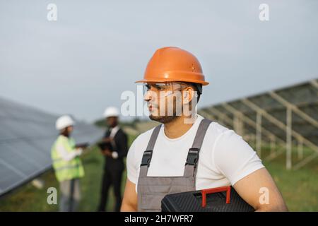 Homme indien dans une combinaison, des lunettes et un casque orange tenant des instruments et regardant de côté en se tenant au milieu de la station solaire.Groupe d'ingénieurs ayant discuté sur les antécédents. Banque D'Images