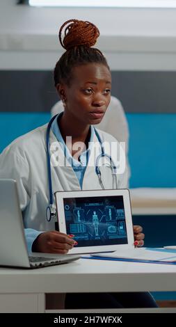 Un patient et un médecin expérimenté regardant une tablette moderne avec analyse du corps humain au bureau de l'armoire. Medic ayant un rendez-vous de contrôle avec l'homme aîné tout en utilisant la technologie dans la chambre d'hôpital. Banque D'Images