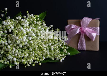 Un bouquet de lys blancs parfumés de la vallée sur fond sombre avec un cadeau en emballage artisanal avec un ruban rose. Banque D'Images