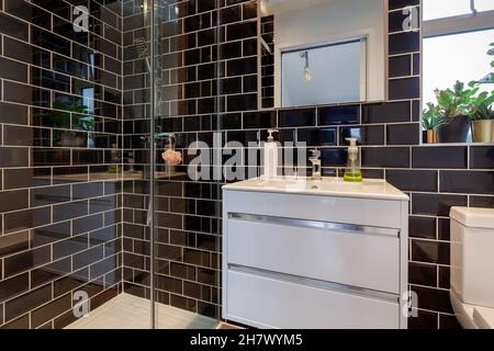 Salle de bain de douche générique avec murs carrelés de noir brillant, cabine de douche en verre, lavabo avec distributeurs de savon et toilettes. Banque D'Images