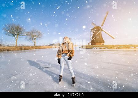 Fille s'amusant sur glace dans un paysage hollandais typique avec moulin à vent.Femme patineuse sur la patinoire en plein air par beau temps de neige.Activités en plein air sur le canal gelé pendant la veille de Noël. Banque D'Images