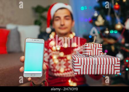Achats de Noël en ligne.Homme achetant des cadeaux du nouvel an à l'aide d'un smartphone.Gars tenant des boîtes-cadeaux et téléphone par arbre de Noël à la maison Banque D'Images