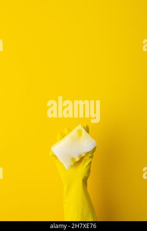 Une main dans un gant de protection jaune en caoutchouc tient une éponge de lavage avec mousse sur un fond jaune Banque D'Images