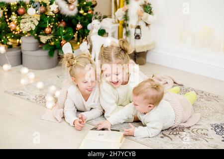 3 filles blondes se trouvent sur le sol près de l'arbre de Noël et lisent un livre. Soirées douillettes avec un livre près de l'arbre.Les amies ont lu un livre ensemble.Chaud r Banque D'Images