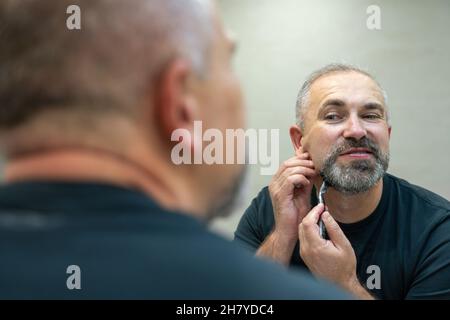 Charmant homme d'âge moyen rasant sa barbe et sa moustache devant un miroir pour paraître plus jeune Banque D'Images