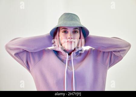 Jeune femme auto-assurée millénaire avec cheveux roses dans le chapeau panama tendance et le sweat à capuche touchant la tête et regardant l'appareil photo sur fond blanc Banque D'Images