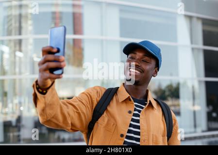 Un homme afro-américain positif avec sac à dos se tenant autoportrait sur un smartphone tout en restant dans la rue près d'un bâtiment moderne dans la ville Banque D'Images