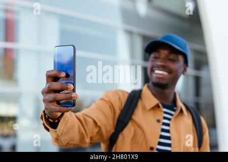 Un homme afro-américain positif avec sac à dos se tenant autoportrait sur un smartphone tout en restant dans la rue près d'un bâtiment moderne dans la ville Banque D'Images