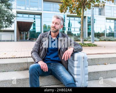 Homme barbu positif soutenu sur une valise tout en étant assis sur des marches de pierre pendant le voyage de travail et en regardant loin Banque D'Images