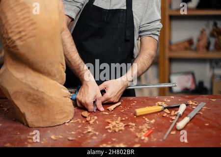 Rognez le menuisier mâle anonyme en tablier à l'aide d'une pince professionnelle tout en sculptant la sculpture en bois à la table avec des burins tout en travaillant dans la menuiserie Banque D'Images