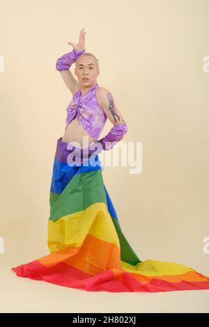 Portrait d'une personne non binaire posant avec un drapeau arc-en-ciel attaché autour de la taille tout en étant sur un fond isolé. Banque D'Images