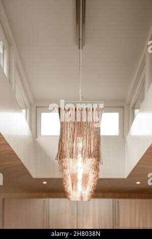 De dessous de lustre lumineux avec de longs éléments décoratifs suspendus plafond blanc contre un mur en bois et une petite fenêtre Banque D'Images