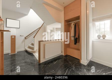 Intérieur d'un hall moderne avec sol carrelé foncé et armoire en bois près des fenêtres placées contre une échelle blanche Banque D'Images