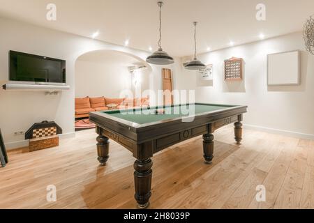 Intérieur d'une salle de billard moderne avec table de billard sous des lustres près de l'espace détente avec canapé et tv au mur Banque D'Images