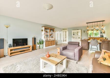 Salon moderne meublé avec canapé confortable et fauteuil autour de la table sur la moquette avec des lampes élégantes au-dessus de la table à manger dans la zone de cuisine i Banque D'Images