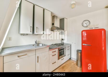 Comptoir avec évier et armoires blanches près du réfrigérateur rouge de style vintage dans la cuisine claire et élégante avec des appareils modernes et horloge dans l'appartement Banque D'Images