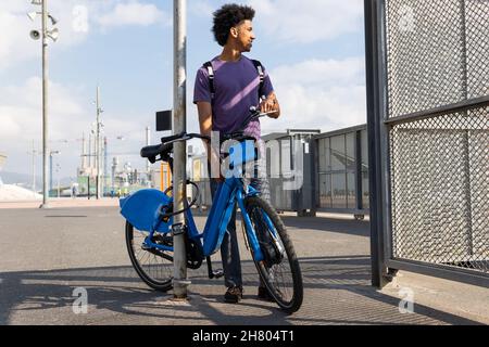 Un jeune homme afro-américain aux cheveux bouclés avec sac à dos debout près du pilier avec vélo et regardant loin avec intérêt Banque D'Images