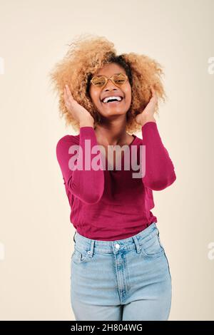 Femme afro touchant ses cheveux et souriant tout en se tenant debout sur un fond isolé. Banque D'Images