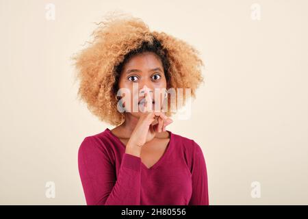 Femme afro surprise faisant un silence ou un geste secret sur un fond isolé. Banque D'Images