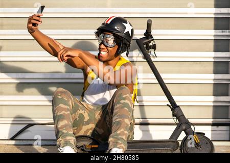 Jeune homme afro-américain positif millénaire en vêtements décontractés et casque de protection assis dans la rue près d'un scooter électrique et prenant un selfie wi Banque D'Images