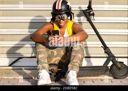 Corps complet de jeunes hommes afro-américains positifs millénaires en vêtements décontractés et casque de protection assis dans la rue près du scooter électrique et de la mesa Banque D'Images