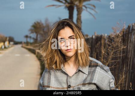 Jeune femme millénaire auto-assurée dans des vêtements élégants et décontractés et des lunettes près de la clôture en bois rustique et regardant l'appareil photo Banque D'Images