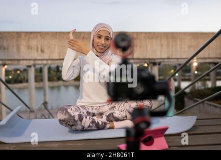 Tout le corps de gaie femme arabe blogger dans hijab tournage vidéo sur la caméra tout en étant assis sur un remblai avec des escaliers contre la rivière Banque D'Images
