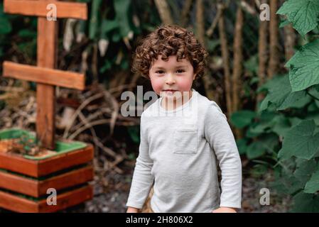 Garçon aux cheveux bouclés debout près du lit de jardin avec des pousses plantées dans la cour et regardant la caméra Banque D'Images