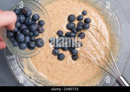 Vue de dessus de crop anonyme personne avec fouet verser les myrtilles dans la pâte dans le bol tout en cuisant des muffins Banque D'Images