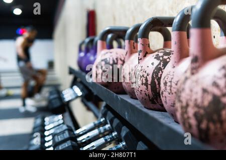 Collection d'haltères et de kettlebells en métal lourd pour soulever la charge placée sur un rack près du mur dans une salle de sport spacieuse avec un sportif sur un arrière-plan flou Banque D'Images