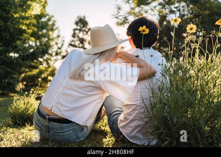 Vue arrière d'hommes et de femmes amoureux méconnus assis ensemble sur un pré herbacé dans la forêt pendant la date romantique Banque D'Images