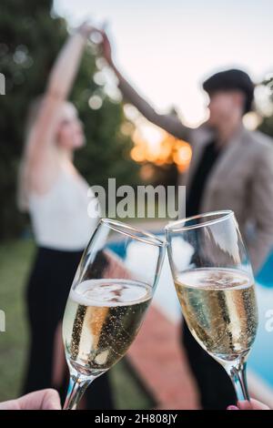 Crop gens toasting avec des verres de boisson alcoolisée sur fond flou de couple romantique avec les bras levés près de la piscine extérieure Banque D'Images