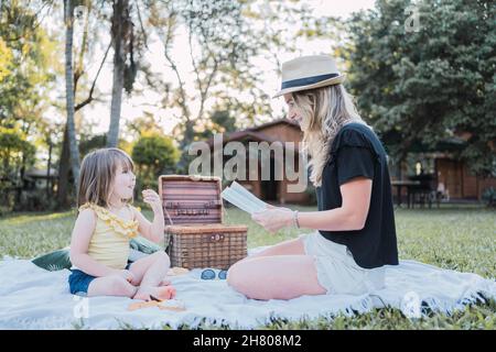 Vue latérale d'une jeune femme souriante avec des cheveux blonds dans des vêtements décontractés et chapeau de paille livre de lecture pour petite fille mignonne tout en étant assis ensemble sur le blanc Banque D'Images