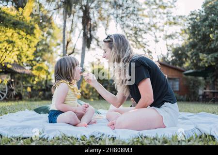 Vue latérale de la jeune mère heureuse dans des vêtements décontractés assis sur une couverture avec des jambes croisées et nourrissant la petite fille mignonne avec un sandwich pendant le pique-nique Banque D'Images