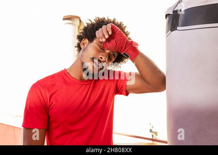 Homme sportif afro-américain épuisé dans un bandage rouge debout près d'un sac de poinçonnage pendant l'entraînement de boxe près d'un bâtiment sur une rue ensoleillée Banque D'Images