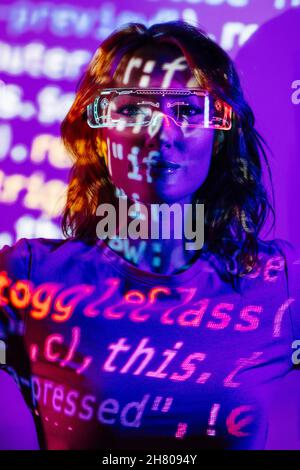 Petit angle de jeune femme dans une tenue décontractée et des lunettes LED debout dans la lumière de néon et regardant l'appareil photo avec code HTML Banque D'Images