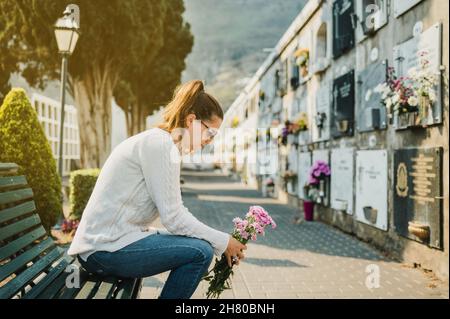 Femme bouleversée assise sur le banc avec des fleurs dans le cimetière Banque D'Images