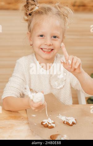 blonde fille peint des biscuits de pain d'épice avec de la crème blanche.L'enfant décorera les biscuits de Noël avec du glaçage sur une table en bois dans la cuisine. Banque D'Images