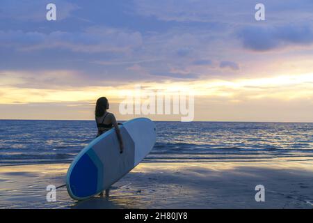 Femme asiatique tenant un panneau supérieur et paddle et marchant sur la plage.En arrière-plan, l'océan et le coucher du soleil.Vue arrière.Surf d'été. Banque D'Images